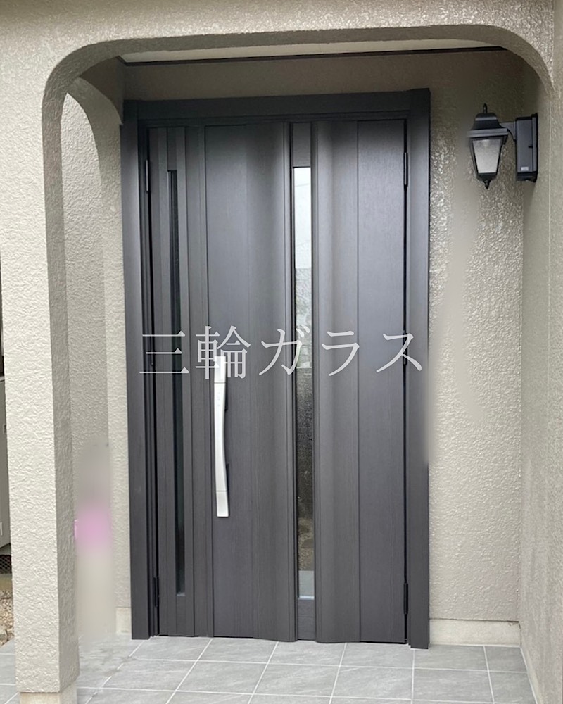 施工が完了したLIXIL「リシェント玄関ドア3」の親子ドア。色は深い木目調のトリノパインで、中央と子扉側に縦長の採光ガラスが入っている。シルバーの長いハンドルが取り付けられ、足元には新しく張り替えられたグレーのタイルが美しく並んでいる。右側の壁には黒いアンティーク調の外灯があり、玄関全体がモダンで落ち着いた雰囲気に仕上がっている。