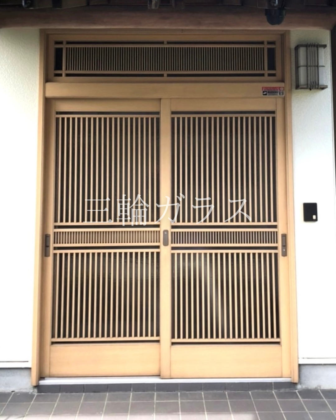 みよし市の和風戸建住宅に設置された、経年劣化した伝統的な木製格子玄関引戸（千本格子）の施工前写真。上部に固定式のランマがあり、和風照明とインターホンが設置されている。建て付けが悪く、鍵が閉まりにくい状態。石畳のポーチ。