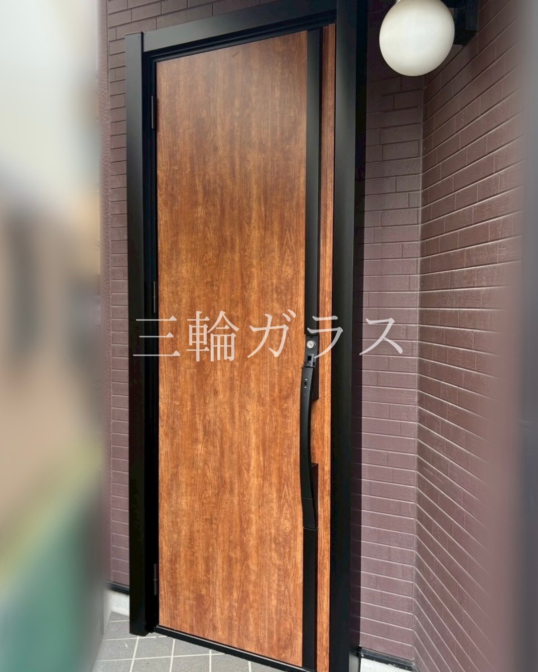 リフォーム後の住宅の玄関ドア。LIXILリシェント（k2断熱仕様）の木目調ハンドダウンチェリーのドアに交換されている。以前の黒い金属枠を利用したカバー工法で、右側に縦長の採光ガラスがあり、モダンな黒いF型ハンドルがついている。外壁のタイルとポーチライトは施工前のまま。