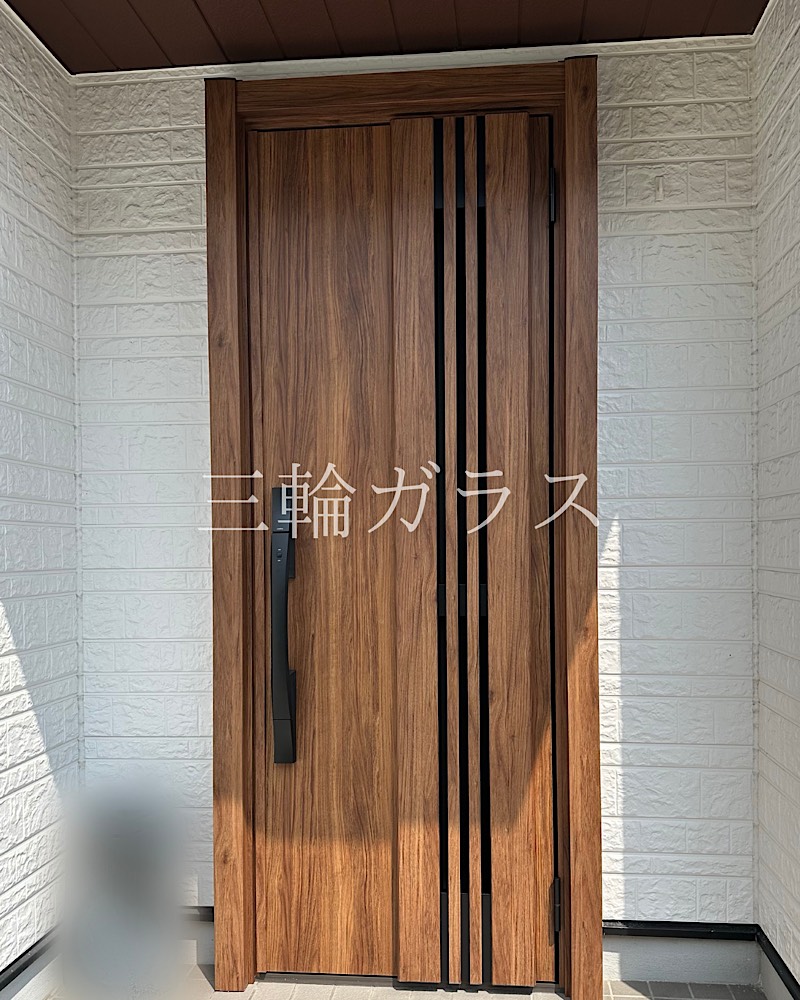 白いレンガ調の外壁に設置された、温かみのあるチーク色の木目調LIXILリシェント玄関ドア3（M83型）。中央に縦のガラススリット、左側に黒いスマートハンドル。中央に「三輪ガラス」の白文字ロゴ。