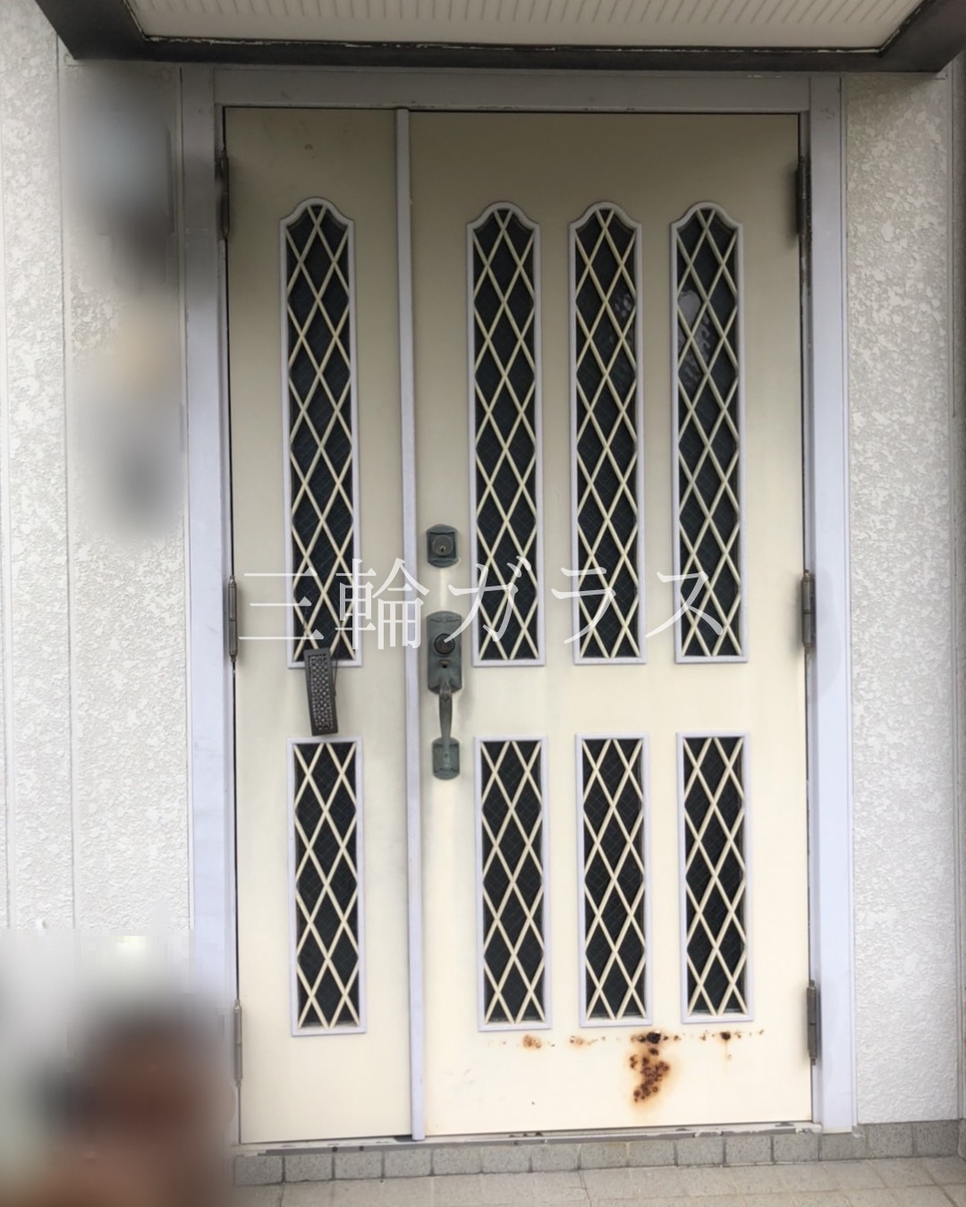 ：錆びと建付けの悪さが目立つ、築33年の白い格子デザインの玄関親子ドア。上部と下部にダイヤ格子のガラス窓があり、古いハンドルと複数の鍵穴が見える。