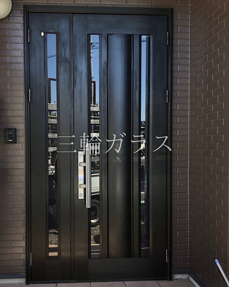 リフォーム前の経年劣化した黒いアルミ製の玄関ドア。ガラス部分に隙間ができ、テープで応急処置がされている様子。